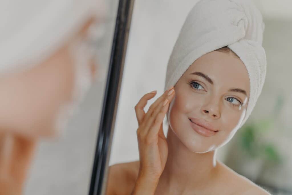 Beautiful woman touches face gently, looks at her reflection in mirror, enjoys softness of skin