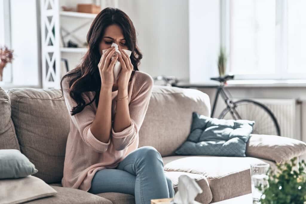 Cold and flu. Sick young woman blowing the nose using tissue paper while sitting on the sofa at home