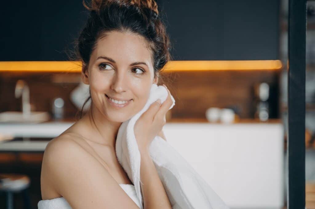 Smiling hispanic girl wiping face by towel enjoy fresh glowing skin after skincare treatment at home