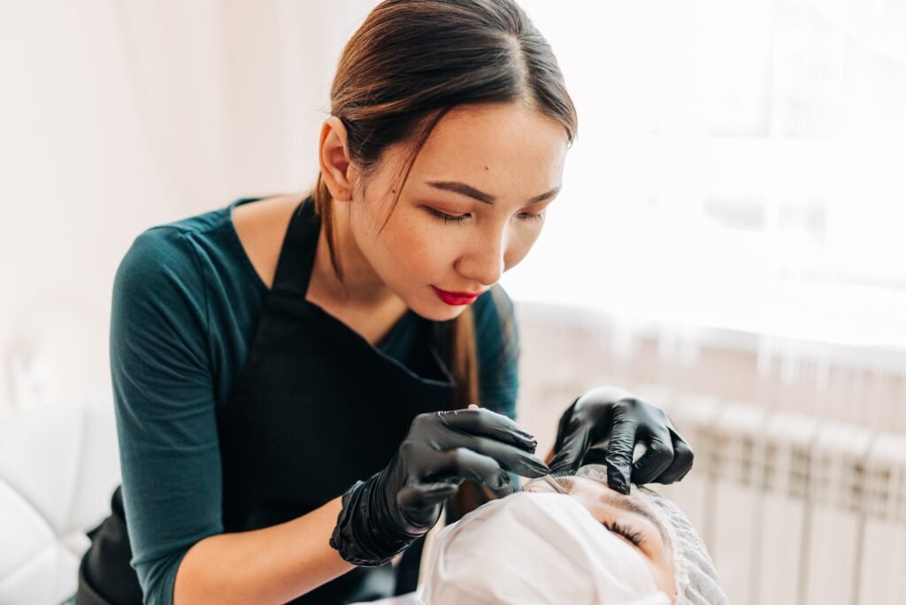 Cosmetologist doing permanent makeup tattooing for brows in her office. Beautiful Asian woman work
