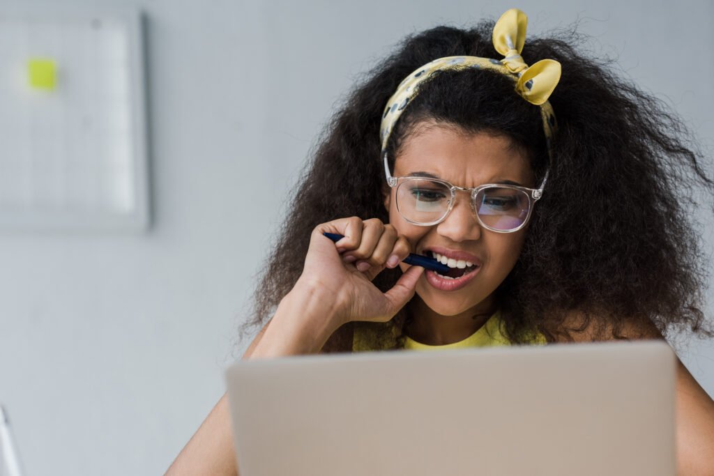 selective focus of emotional african american girl biting pen while looking at laptop